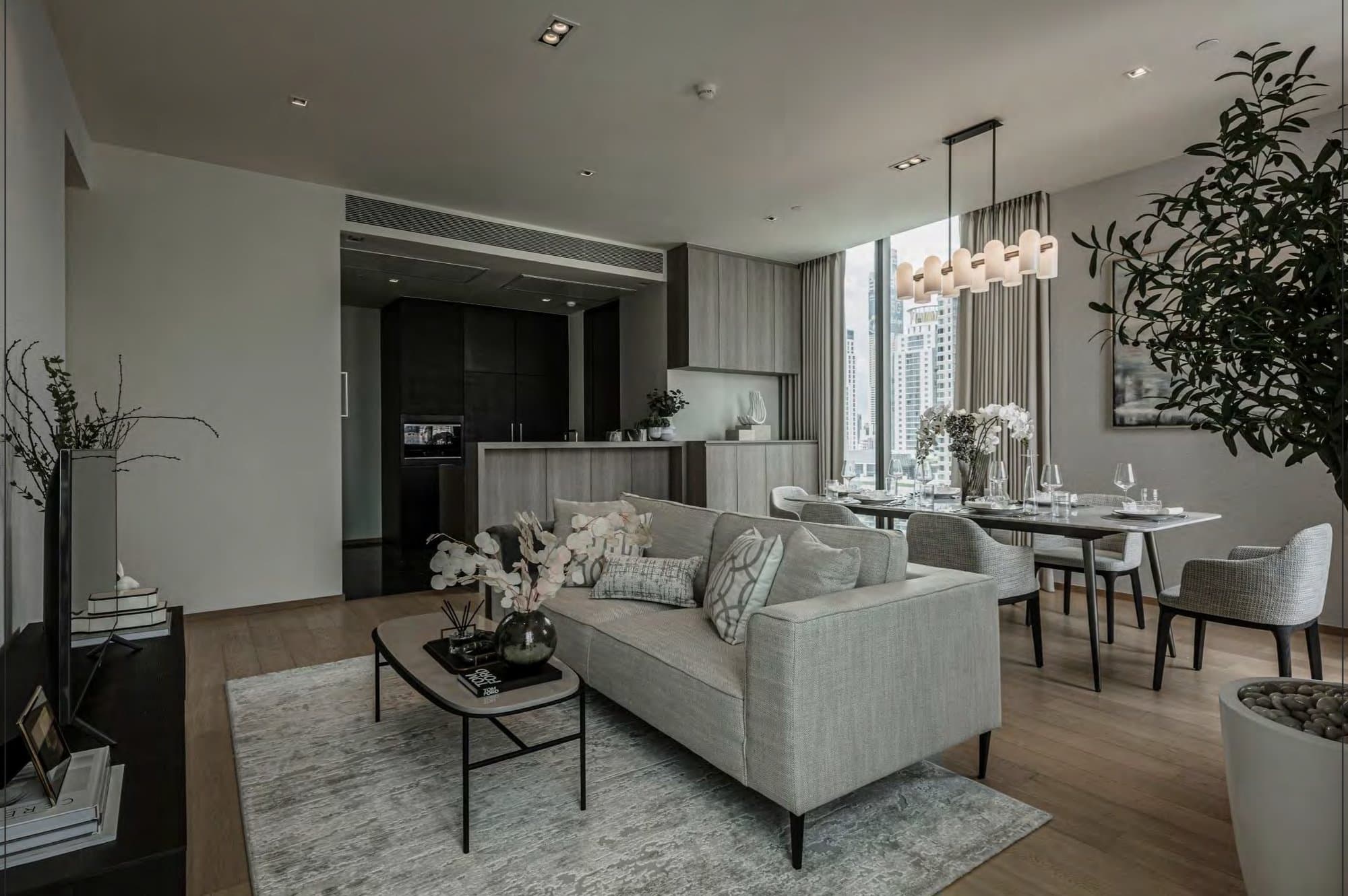 Living and dining area with pale linen sofa, oval coffee table on a soft rug, open kitchen behind and an olive tree by the glazing