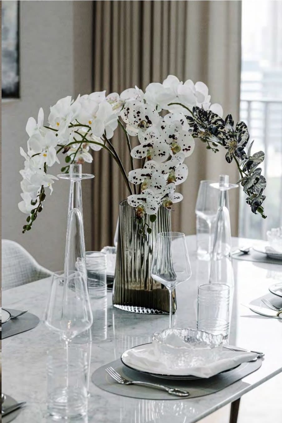 Tablescape detail of white orchids in a fluted smoke-glass vase, slender wine glasses and a gold-rimmed dish on white marble