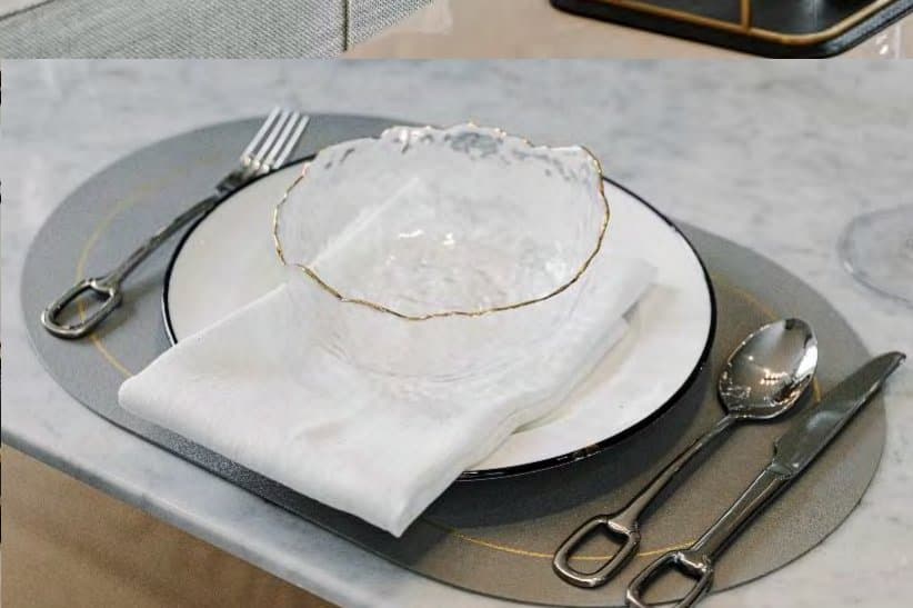 Place setting close-up with a gold-edged textured glass bowl, folded linen napkin, charger plate and polished cutlery on white marble