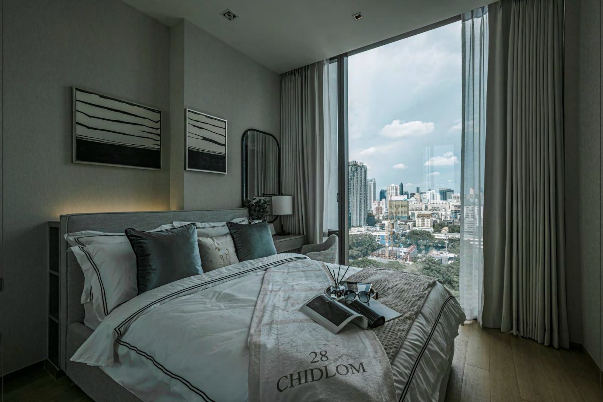 Bedroom in muted greys with layered white linens, teal cushions, framed monochrome art and a wide window over the leafy Bangkok skyline