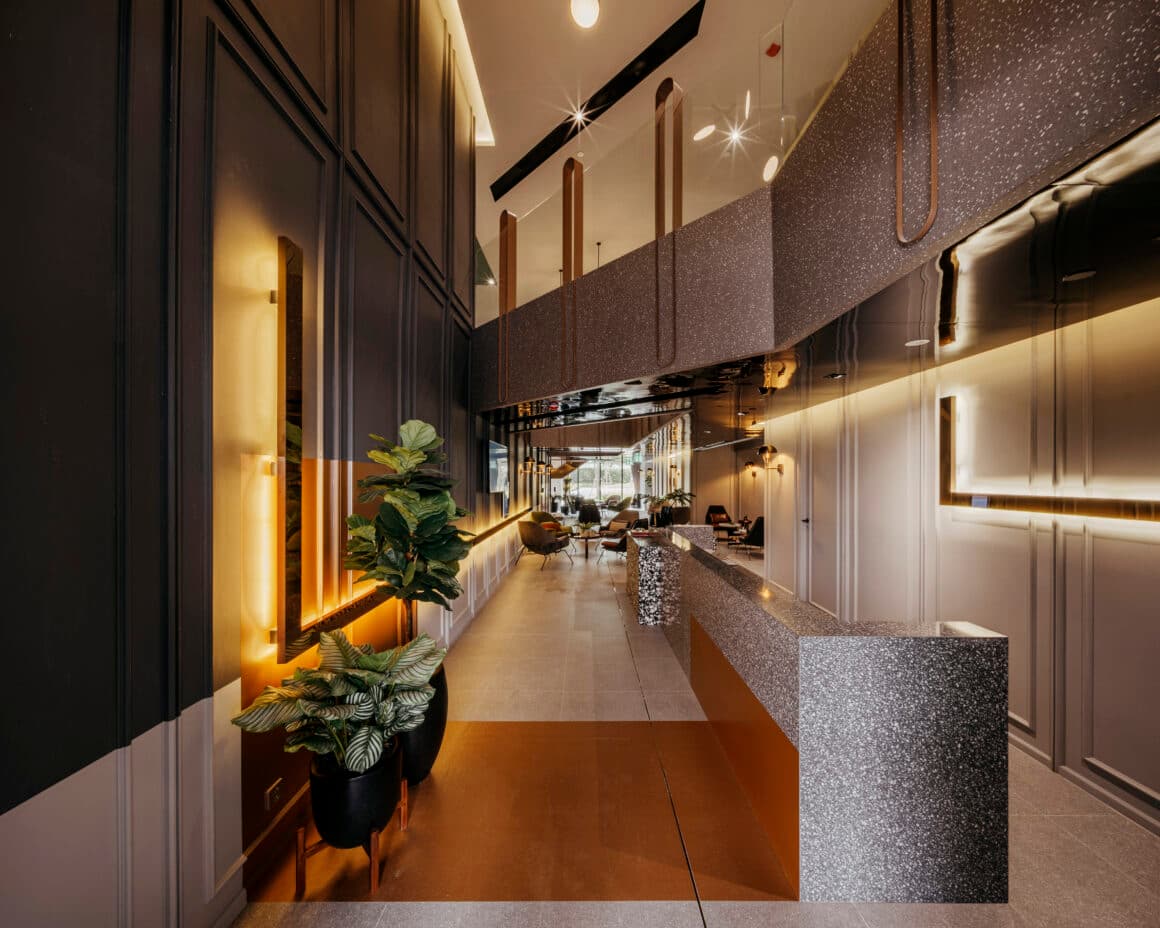 Lobby entrance with grey terrazzo reception counter, dark panelled walls, warm backlit sconces, potted plants and copper flooring