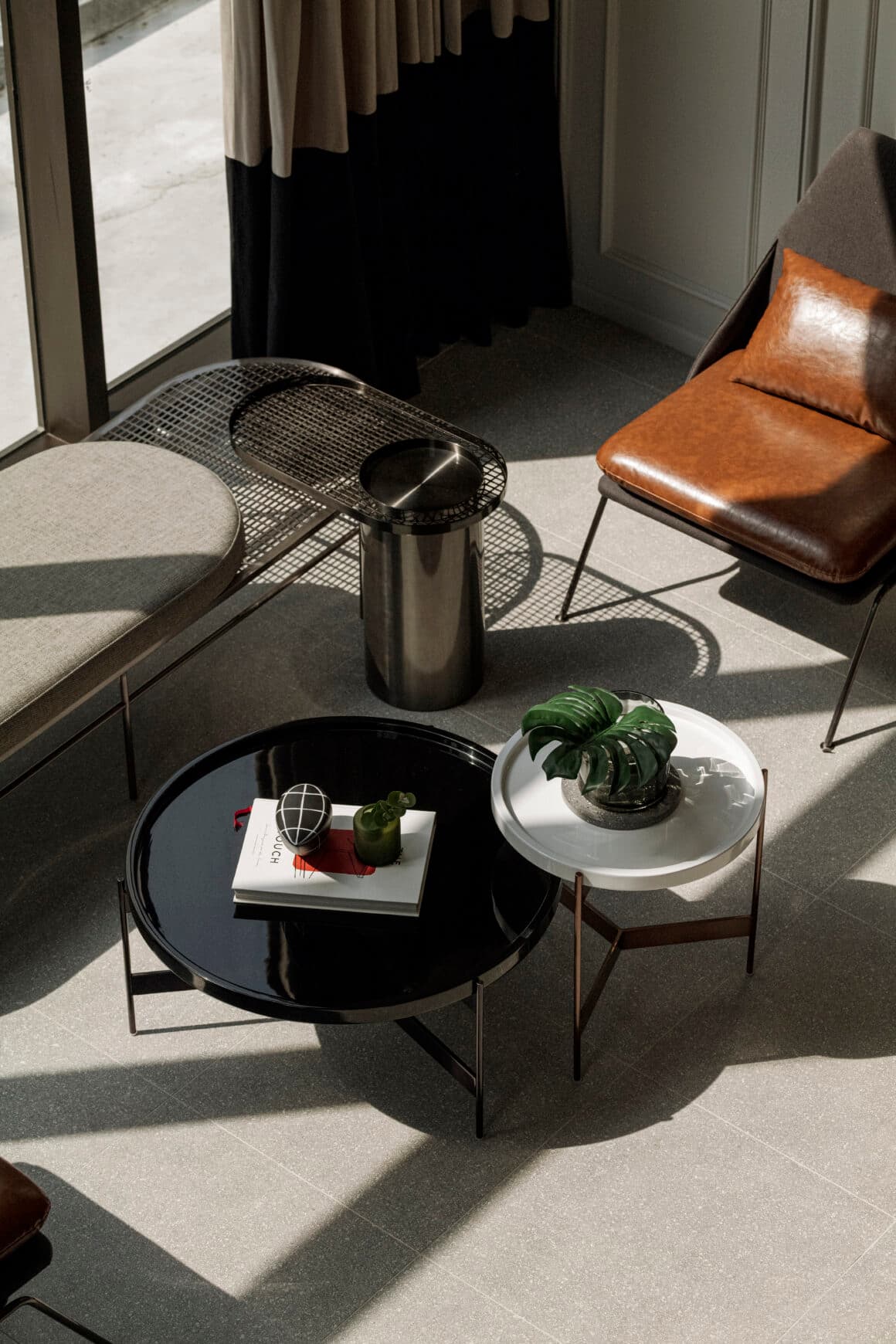 Sunlit lounge corner seen from above: nesting black and white coffee tables, tan leather armchair and grey upholstered bench