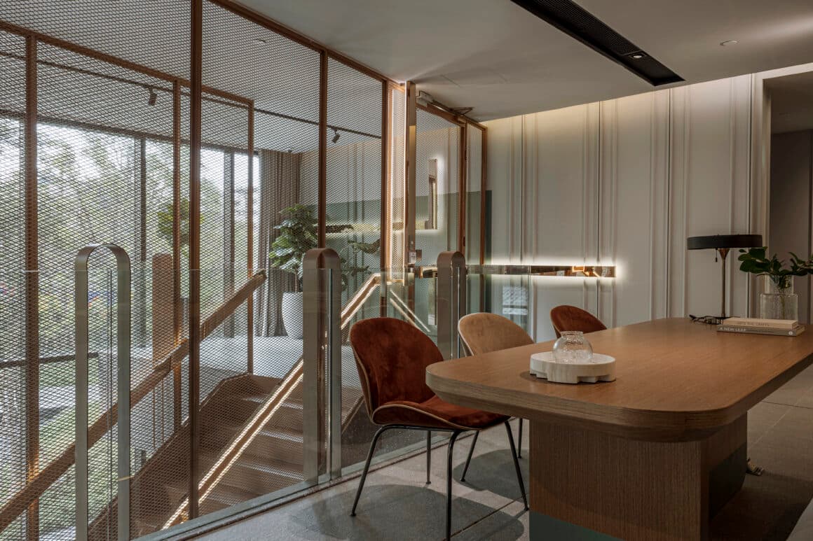 Meeting corner with oval oak table, rust velvet chairs and black lamp beside a bronze mesh and glass stairwell