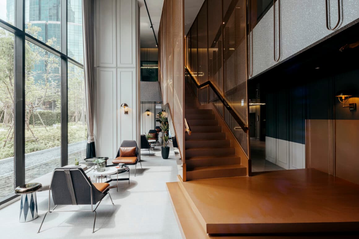 Double-height co-working lounge with grey armchairs, leather cushions, tall garden-facing windows and caramel-toned staircase with mesh screen
