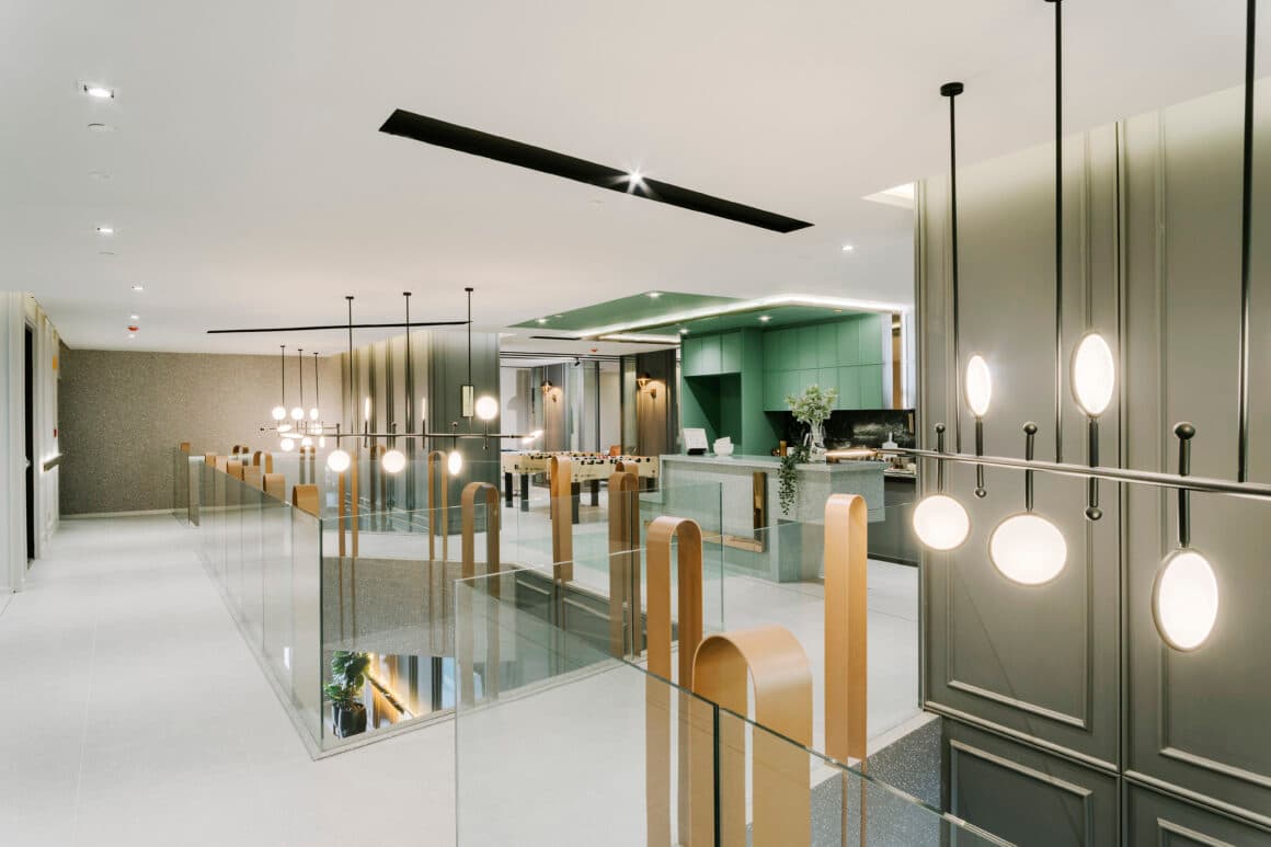 Mezzanine games lounge with glass balustrades, brass loop details, globe pendant lights, sage-green pantry cabinets and foosball table