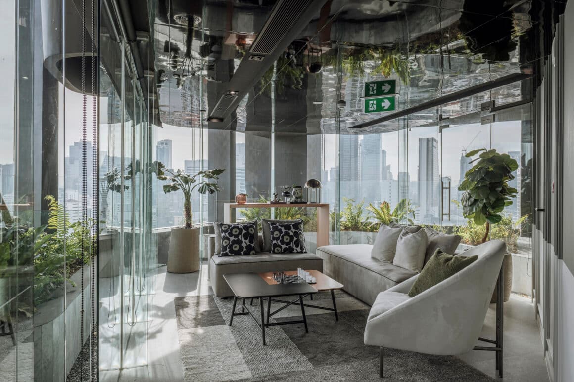 Glass-walled sky lounge with cream sofas, patterned cushions, lush plants, mirrored ceiling and panoramic Bangkok skyline views