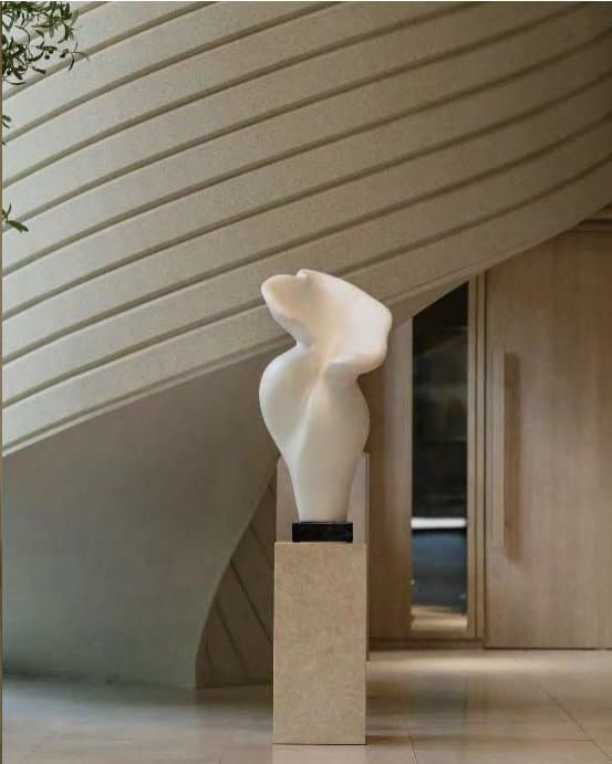 A smooth alabaster-white abstract sculpture on a stone plinth, set against warm ribbed plaster walls and pale timber doors.