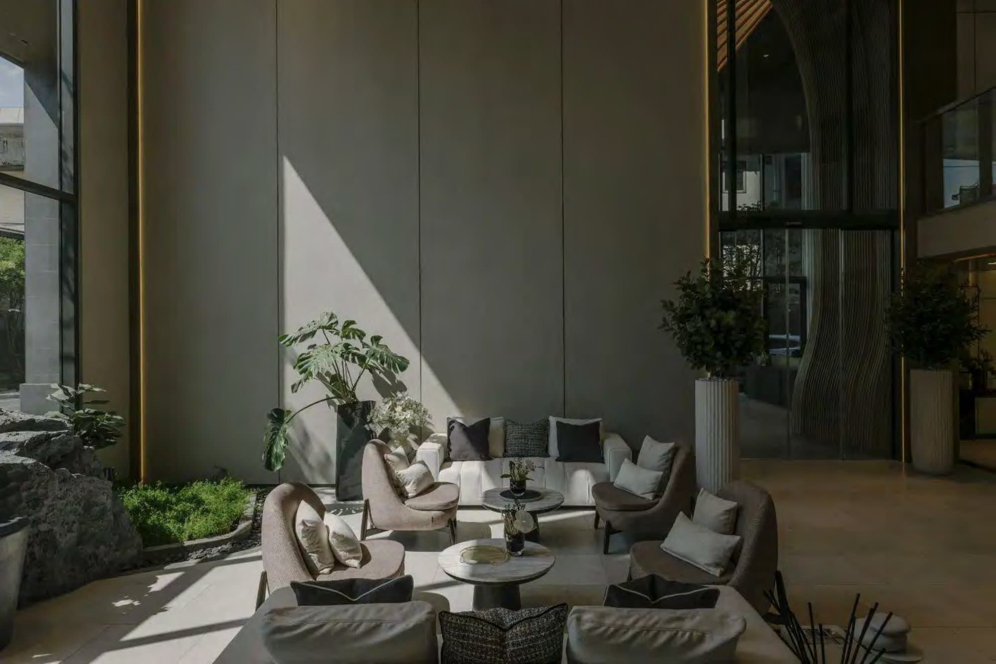 Soaring grey-panelled lounge where bouclé armchairs gather round marble tables, a monstera and indoor garden softening full-height glazing.
