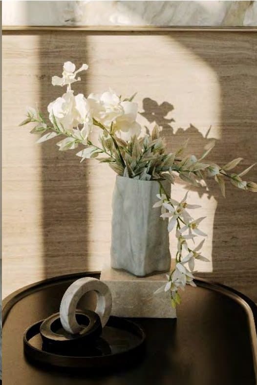 White orchids and slender foliage in a pale sculptural vase on a black tray, sunlight throwing soft shadows across travertine.