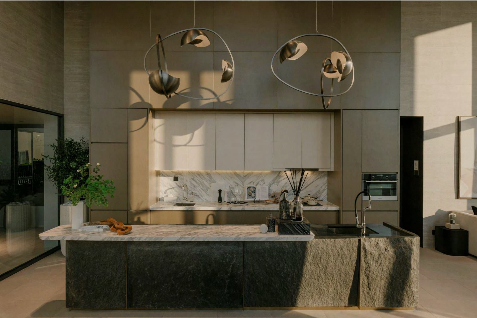 Show-unit kitchen with a rough-hewn dark stone island, marble worktop, marble splashback and two sculptural bronze ring pendants overhead.