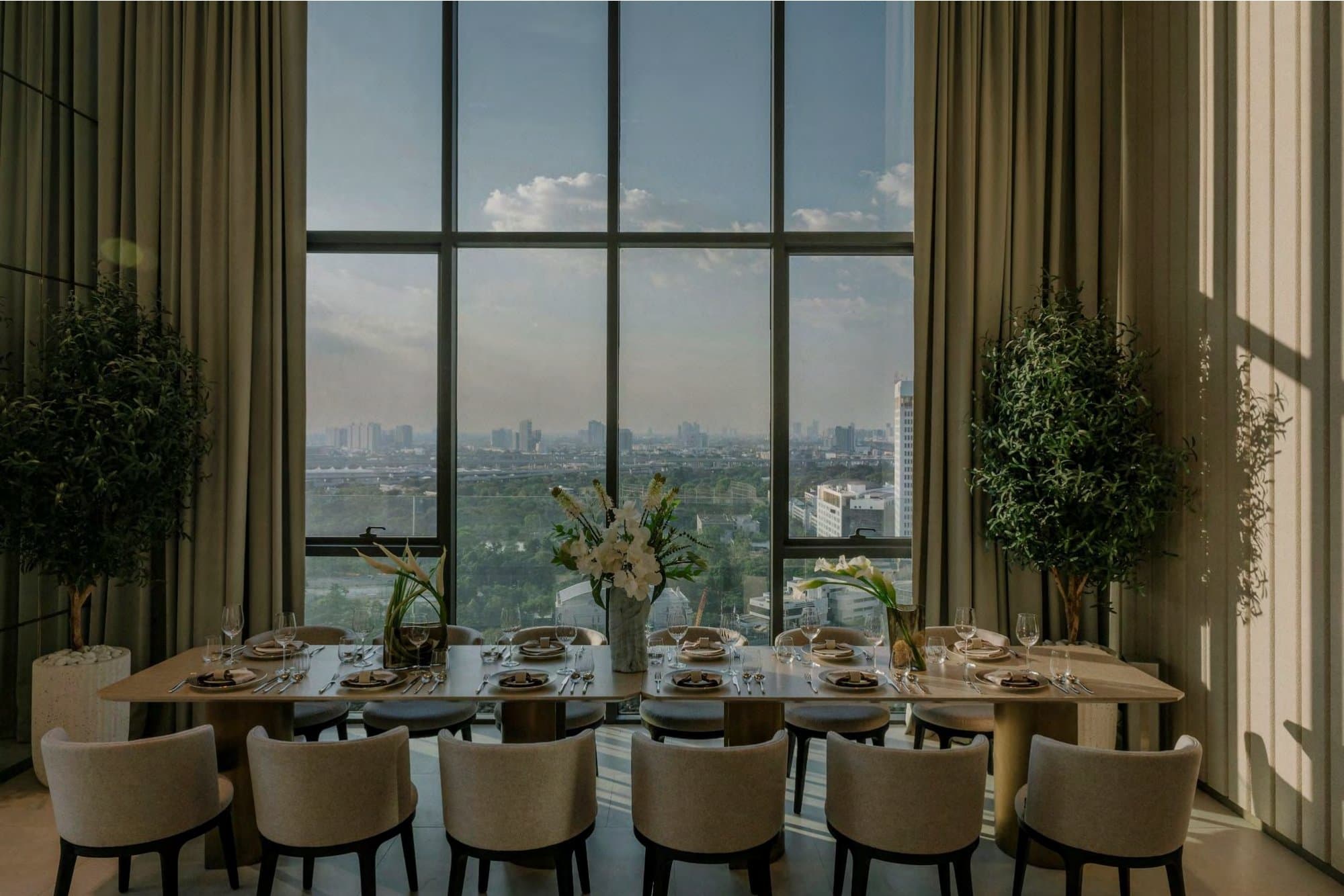Dining table set for twelve before a double-height window framing a panoramic Bangkok skyline, flanked by olive topiaries and linen curtains.