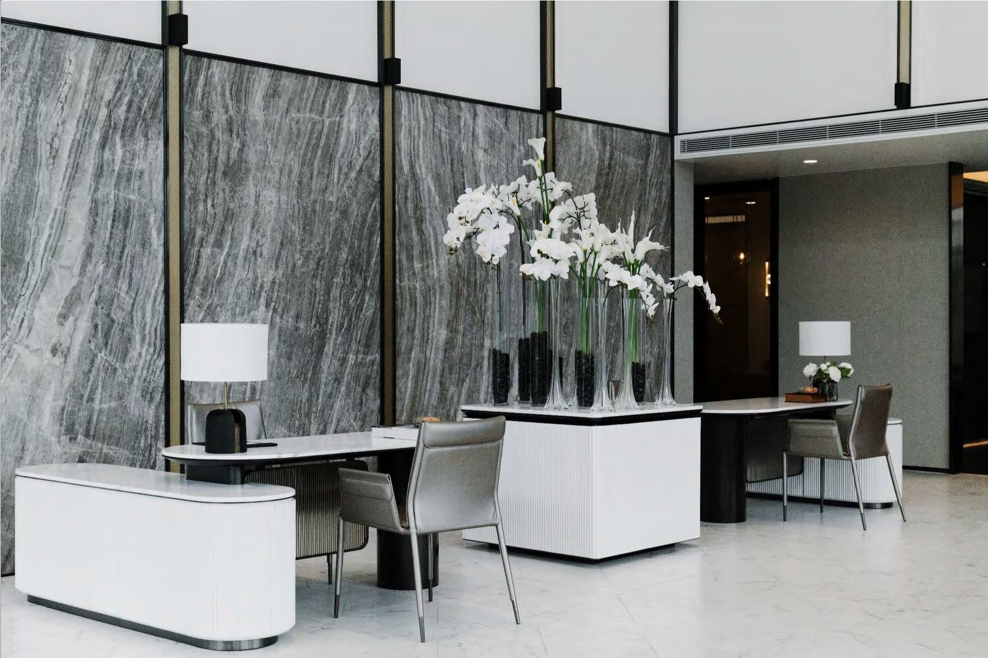 Reception desks against a grey marble wall, white orchids in tall glass vases, table lamp and grey leather chairs.