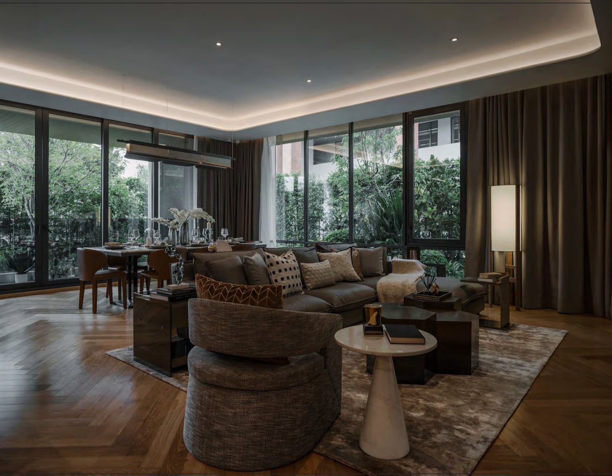 Living room at dusk with a brown leather sectional, round bouclé chair, marble side table and glazing onto dense greenery.