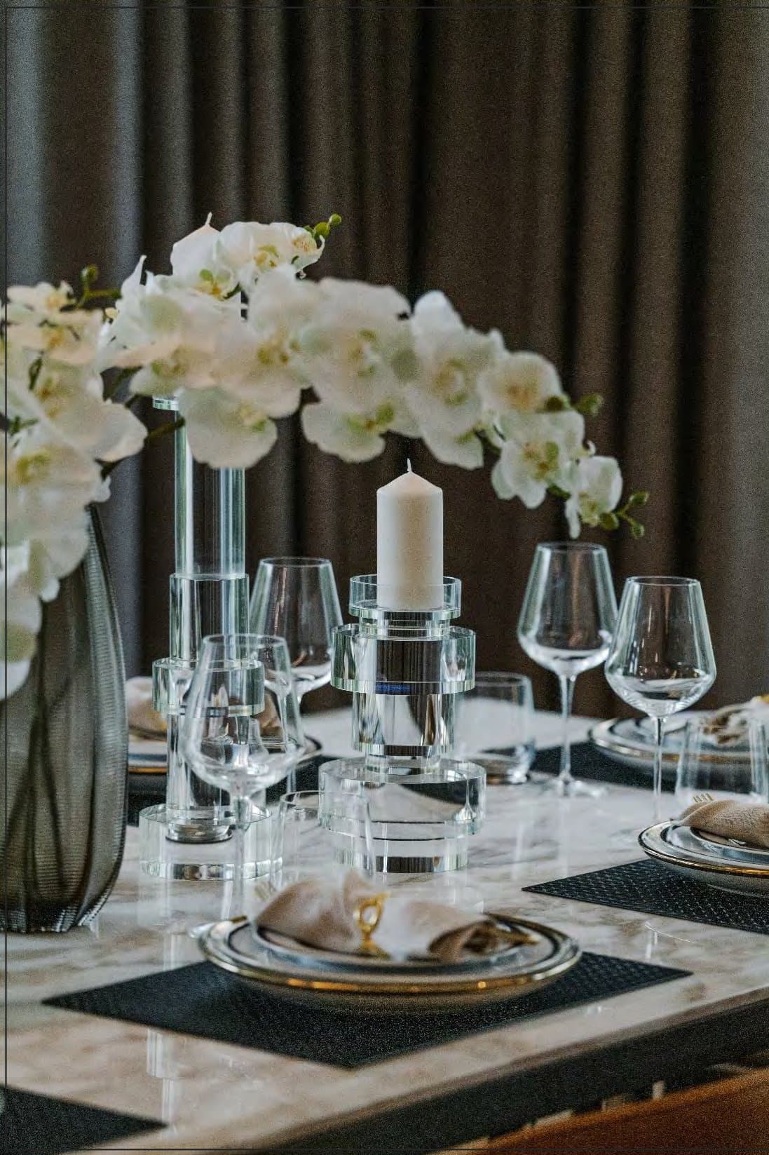 Table setting detail: marble surface, gold-rimmed plates, crystal candle holder, clear wine glasses and a spray of white orchids