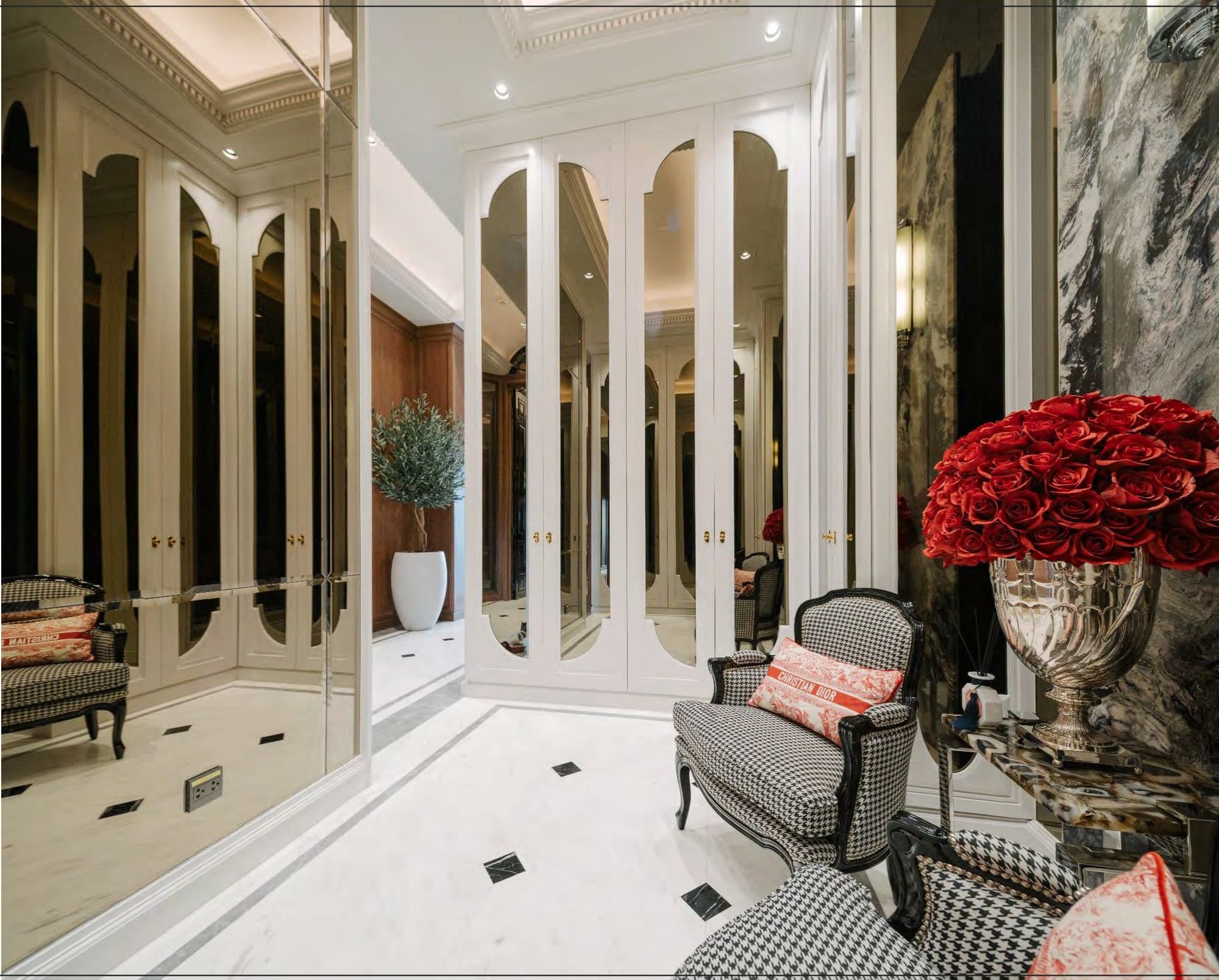 White dressing room lined with arched mirrored wardrobes, black-and-white houndstooth Louis chairs and a silver urn brimming with red roses.