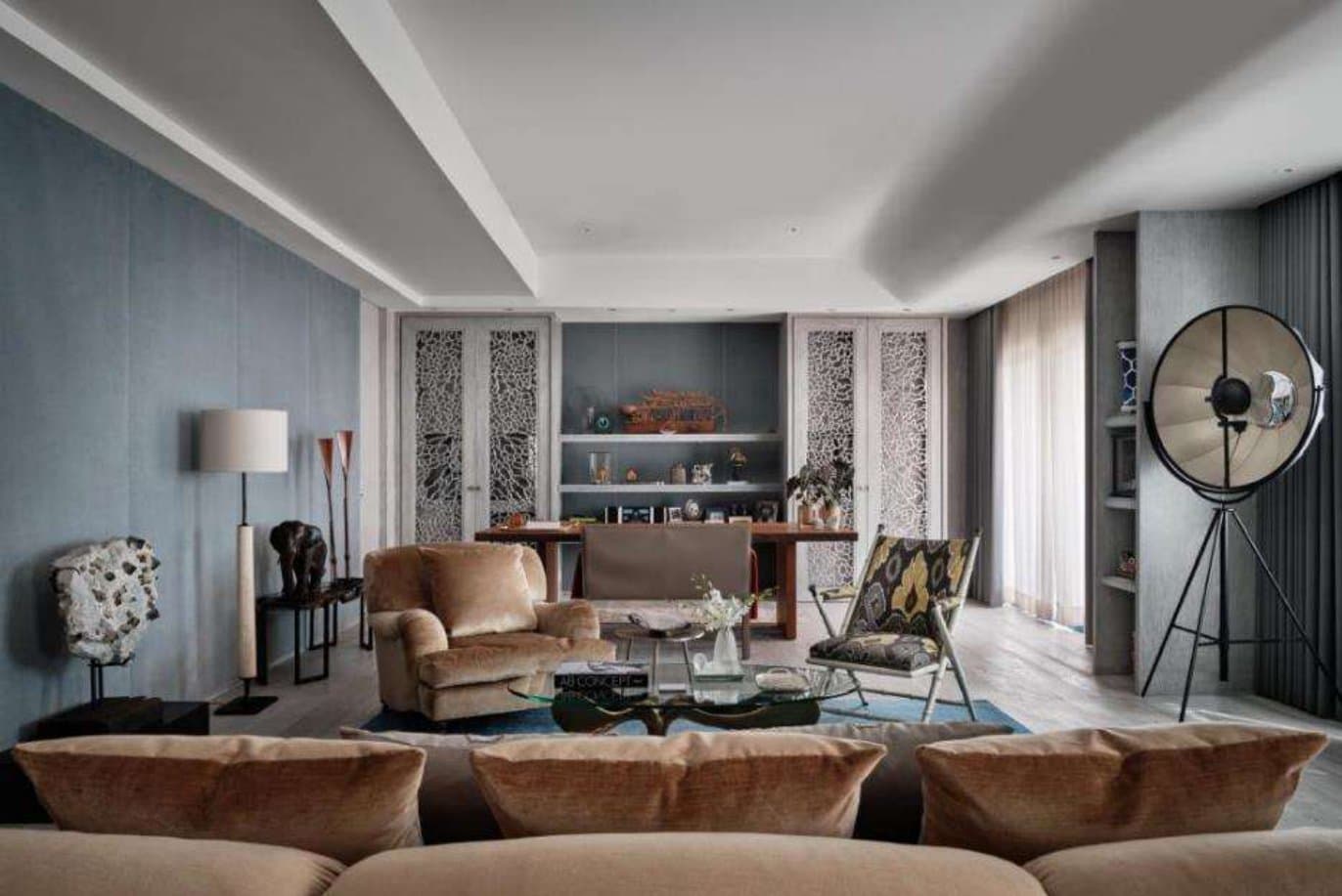 Wide living room view with camel velvet seating, a tripod fan-lamp, sheer curtains and lattice cabinet doors framing lit display shelves.