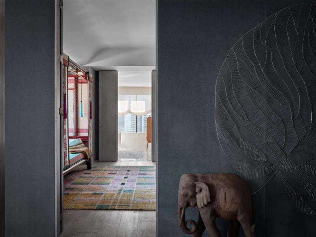Charcoal-walled threshold with a carved wooden elephant and incised leaf relief, opening to a bedroom with plaid rug and city view.