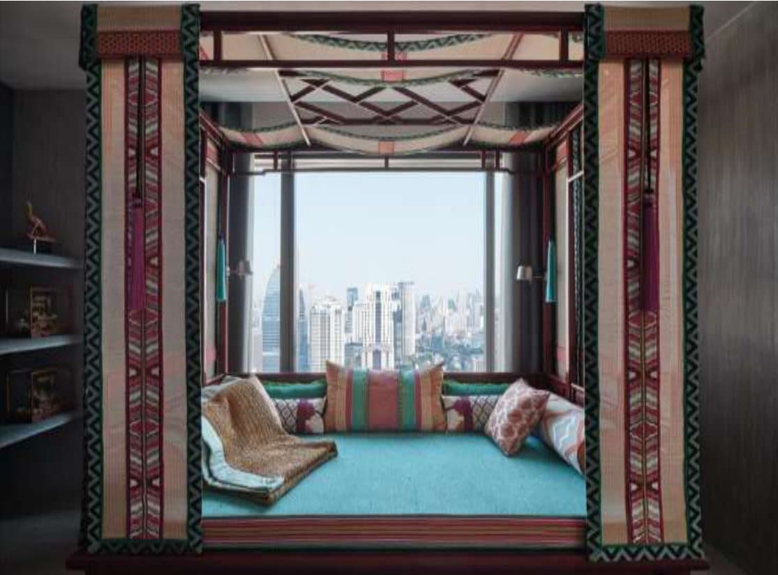 Canopied daybed nook framed in tribal-patterned textiles, turquoise cushions and striped pillows, set against floor-to-ceiling windows overlooking the Bangkok skyline.