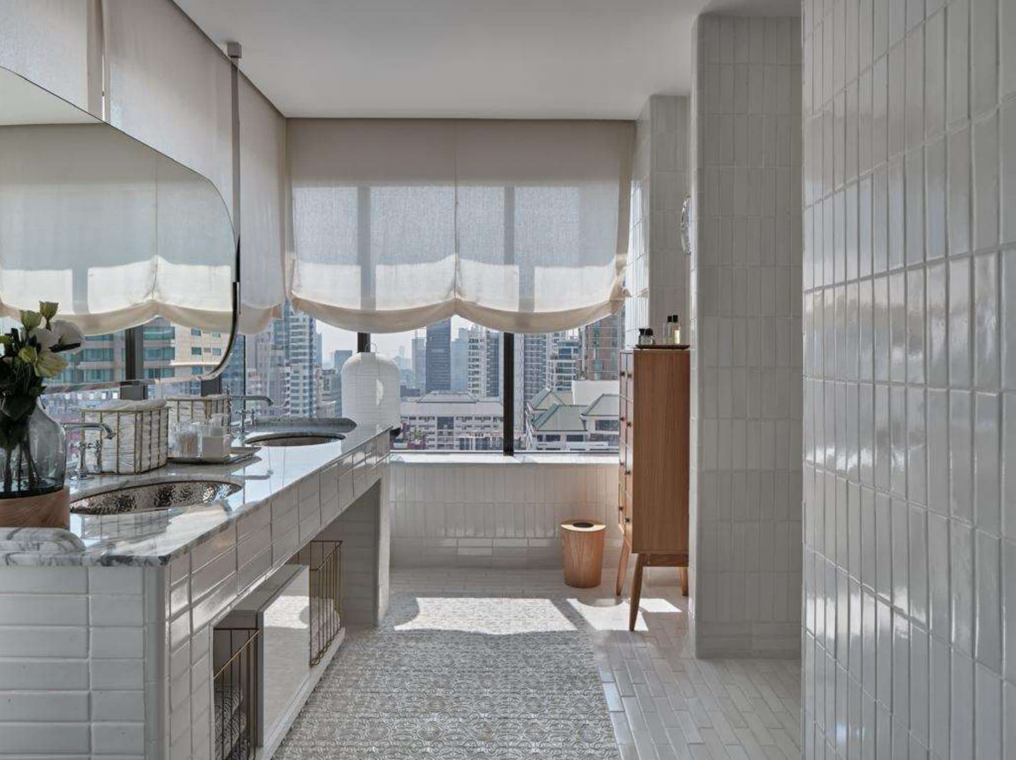 Sunlit master bathroom with white tiled double vanity, hammered-silver basins, marble counter, sheer roman blinds and a high-rise city outlook.