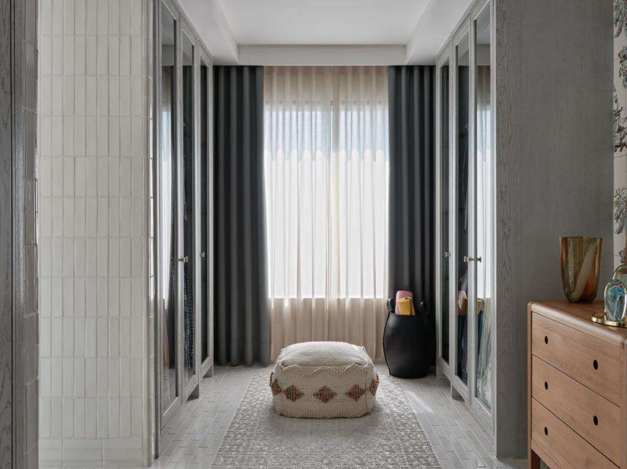 Dressing room lined with mirrored wardrobes, grey drapes over sheers, a woven pouf on a patterned rug and a timber dresser.