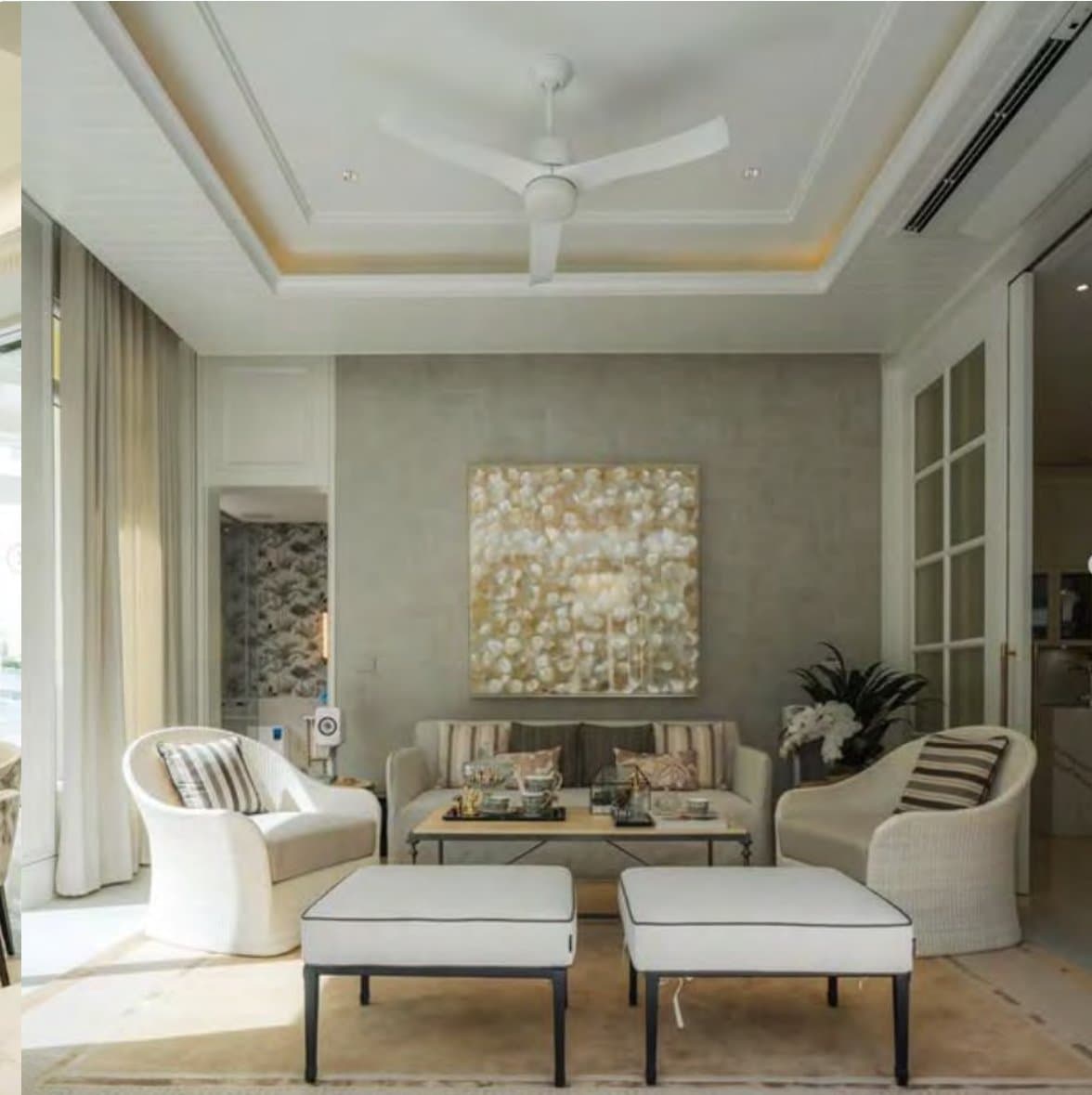 Family sitting room under a coffered ceiling and white fan, cream tub chairs and a striped settee facing a textured pearl-and-gold canvas on a grey wall