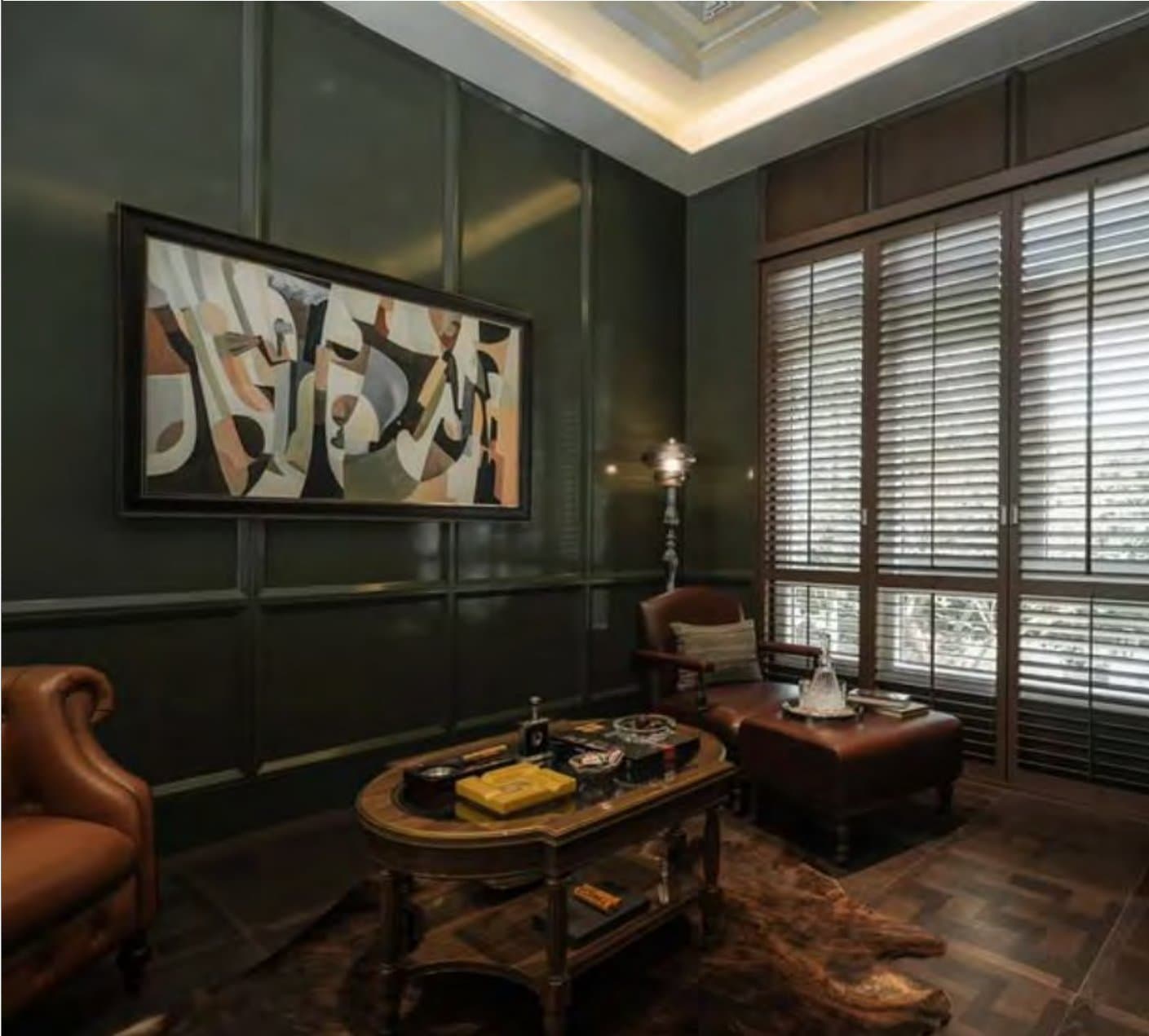 Cigar lounge in forest-green panelling with a tan leather wing chair, oval timber table, large abstract canvas and shuttered floor-to-ceiling windows