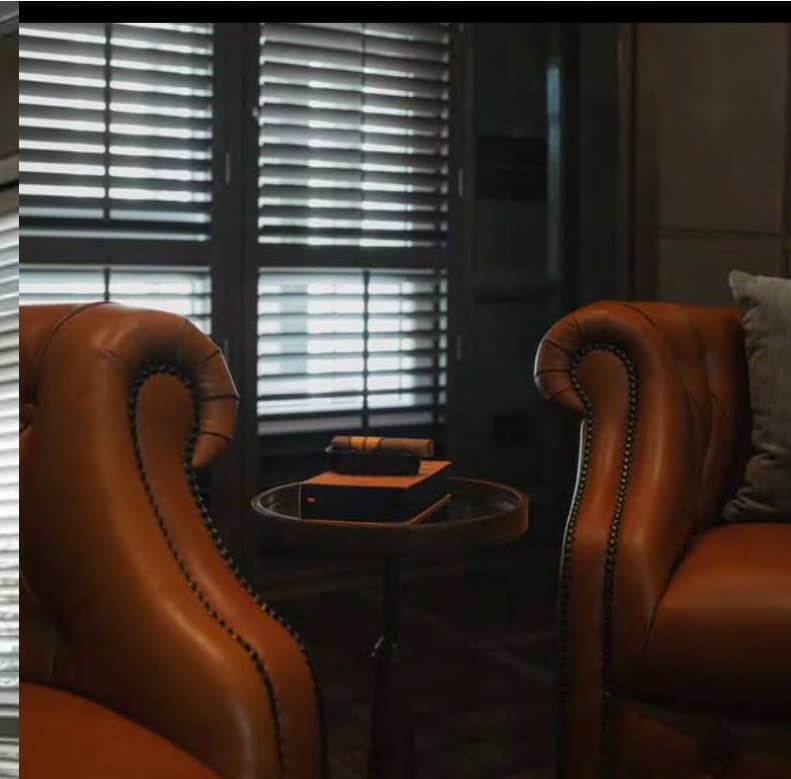 Two tan leather Chesterfield wing chairs with nailhead trim face across a small round table set with a cigar box, plantation shutters behind