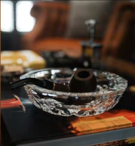 Cut-crystal ashtray cradling a smoking pipe on a lacquered table, a leather wing chair blurred in the warm background