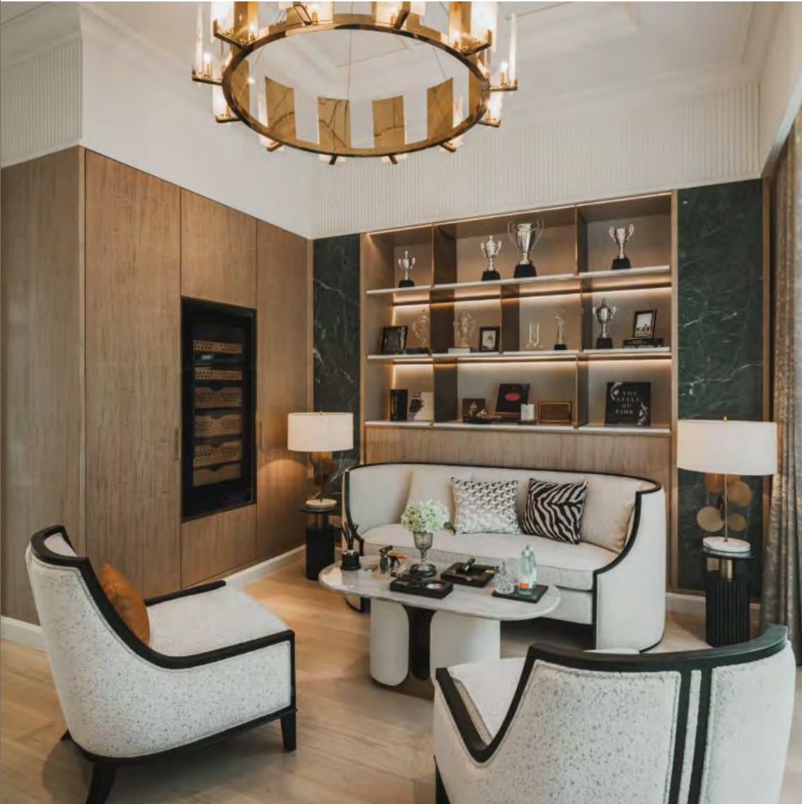Cigar lounge with wood-panelled walls, backlit shelves of trophies, a curved cream sofa and paired black-and-white bouclé armchairs.