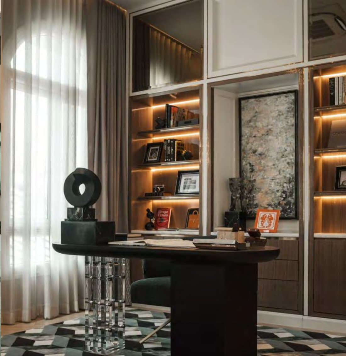 Home-office corner with a dark oval desk on crystal legs, backlit bookshelves, framed abstract art and a geometric green rug.