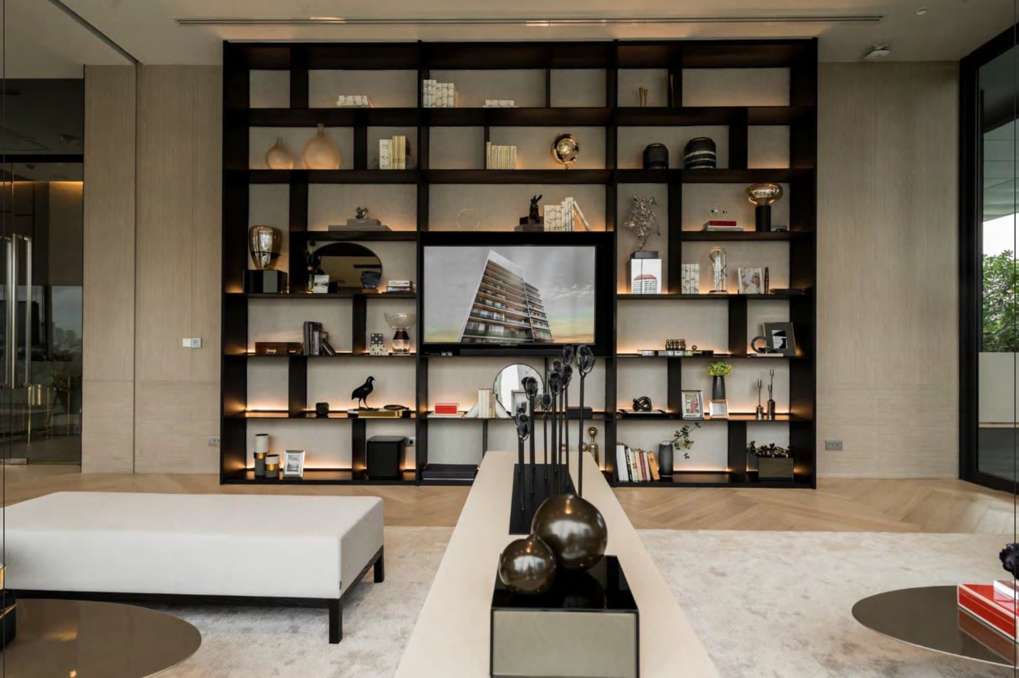 Full-height black display wall of backlit open shelving holds books, brass vessels and objets around a central screen above pale parquet