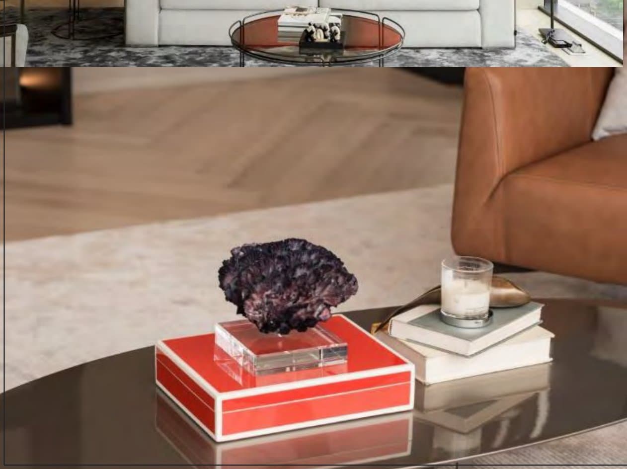 Styling detail of a coral lacquer tray holding a violet geode on a bronze table, beside stacked books, a lit candle and tan leather chair