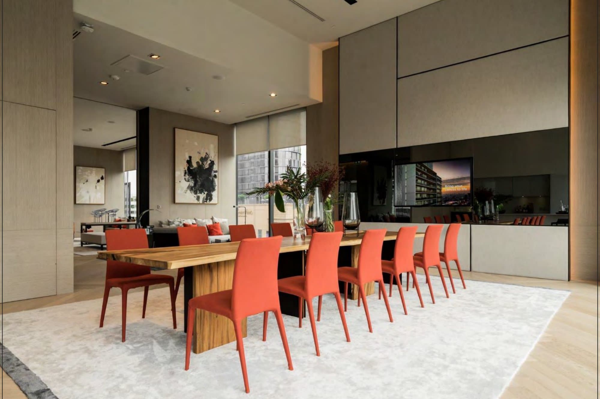 Long live-edge timber dining table lined with coral upholstered chairs on a pale rug, ink artworks and city views behind