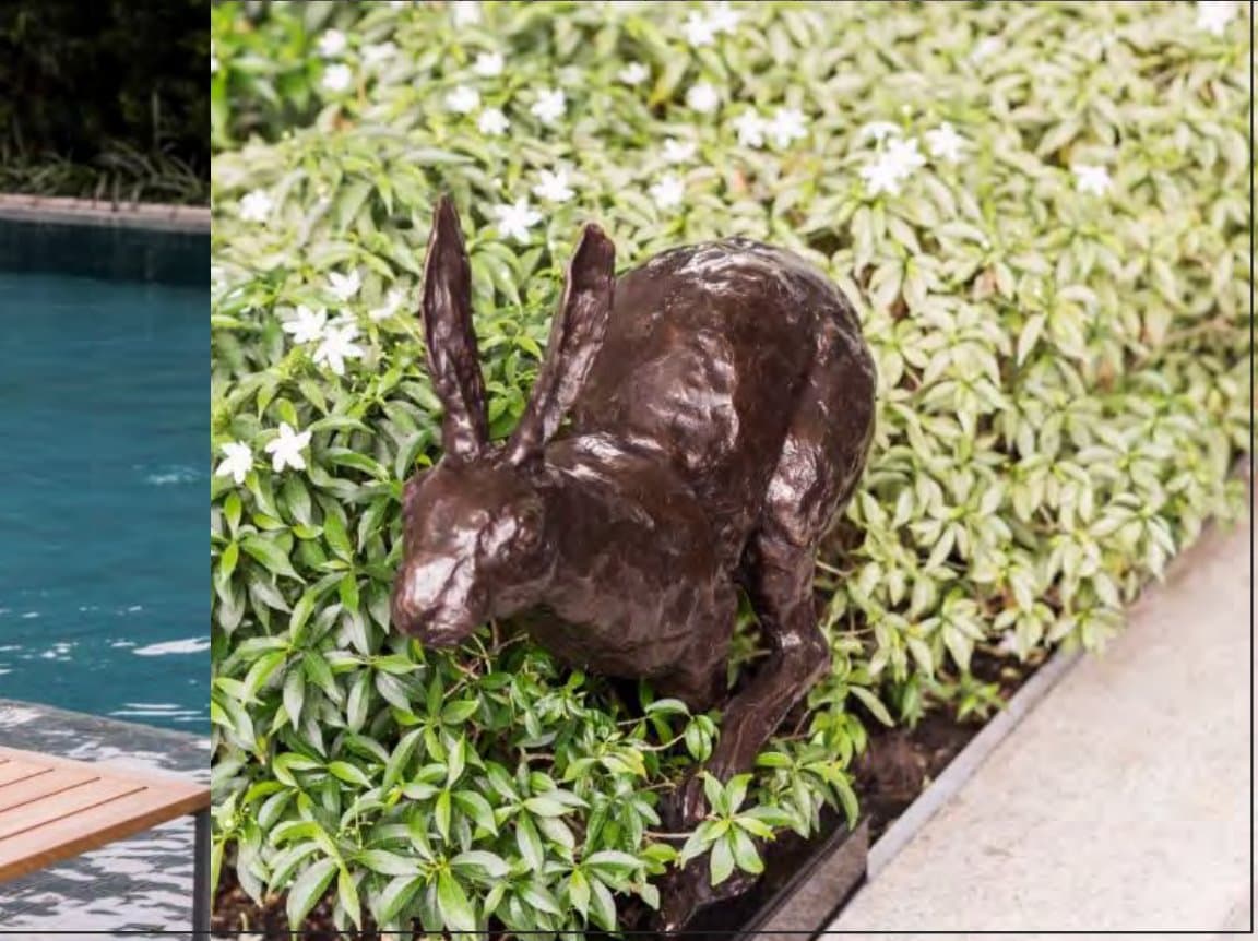 Bronze hare sculpture crouches among glossy leaves and small white blooms at the planted edge of a turquoise swimming pool