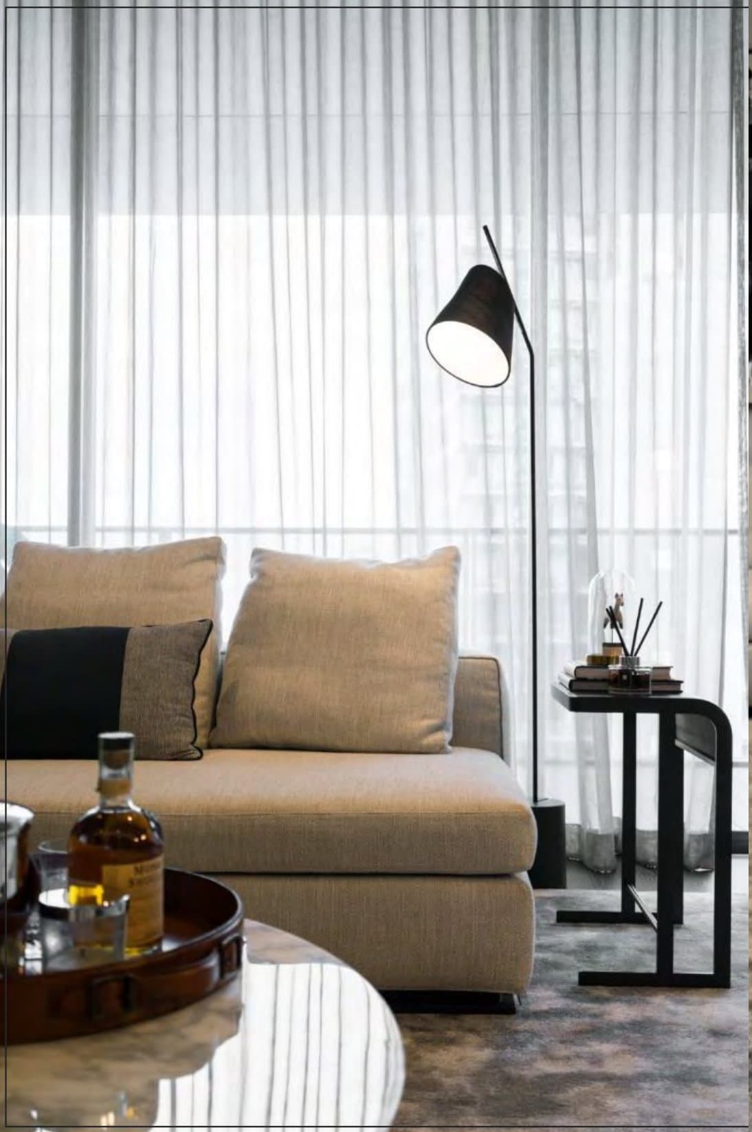 Quiet corner where a taupe sofa meets a slim black arc lamp and side table, sheer curtains diffusing daylight, whisky tray in the foreground