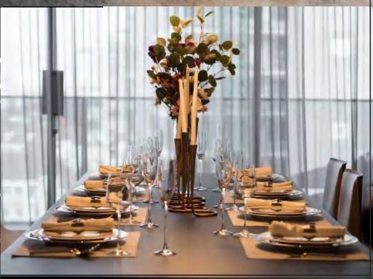 Dressed dining table set with charcoal plates, gold-ringed napkins, tall brass candlesticks and an autumnal eucalyptus centrepiece before sheer curtains