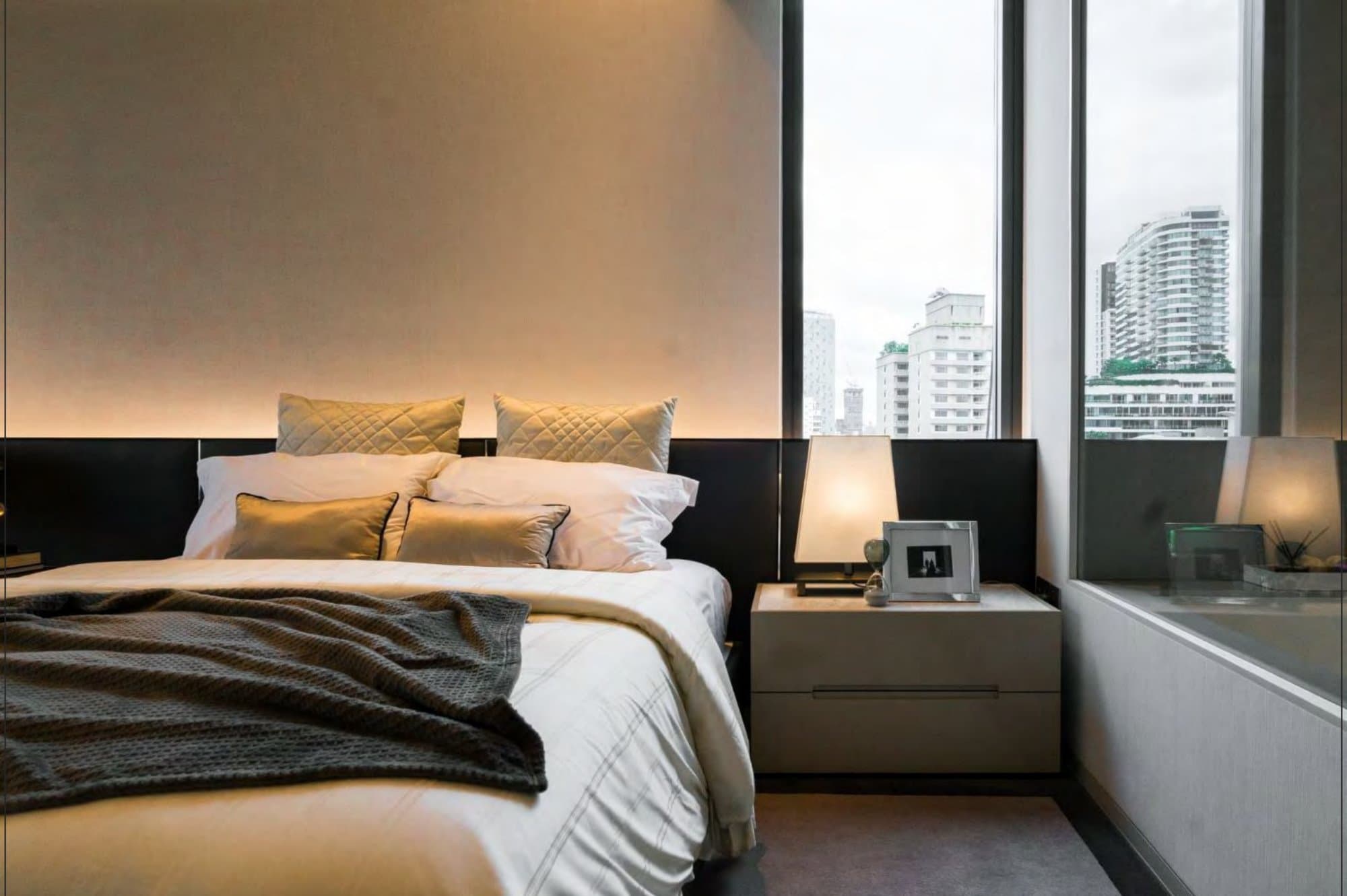Warm bedroom with a backlit upholstered headboard, layered white linens, gold quilted cushions and corner windows overlooking the city