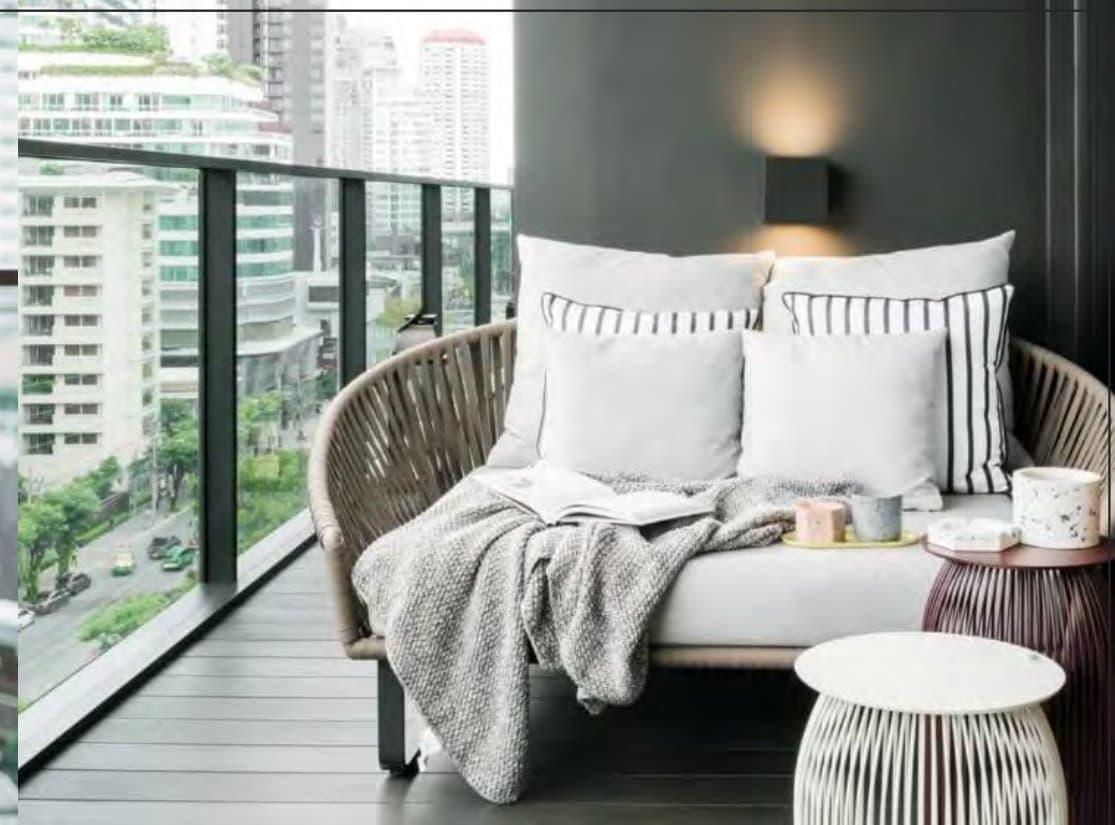 Balcony loveseat of woven cord piled with white and striped cushions and a knit throw, overlooking mid-rise buildings and street greenery