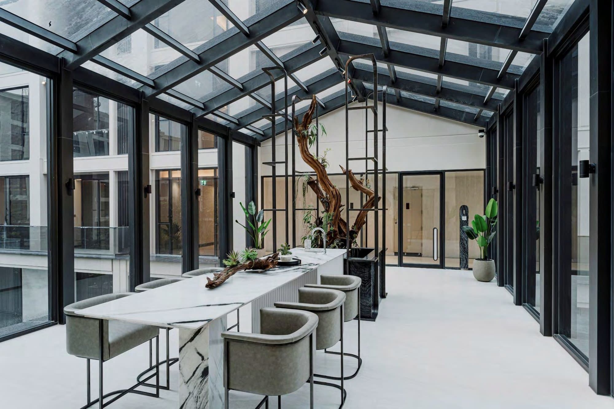 Glass-roofed conservatory lounge under a black steel gable, with a veined marble bar, leather stools and a sculptural driftwood centrepiece.