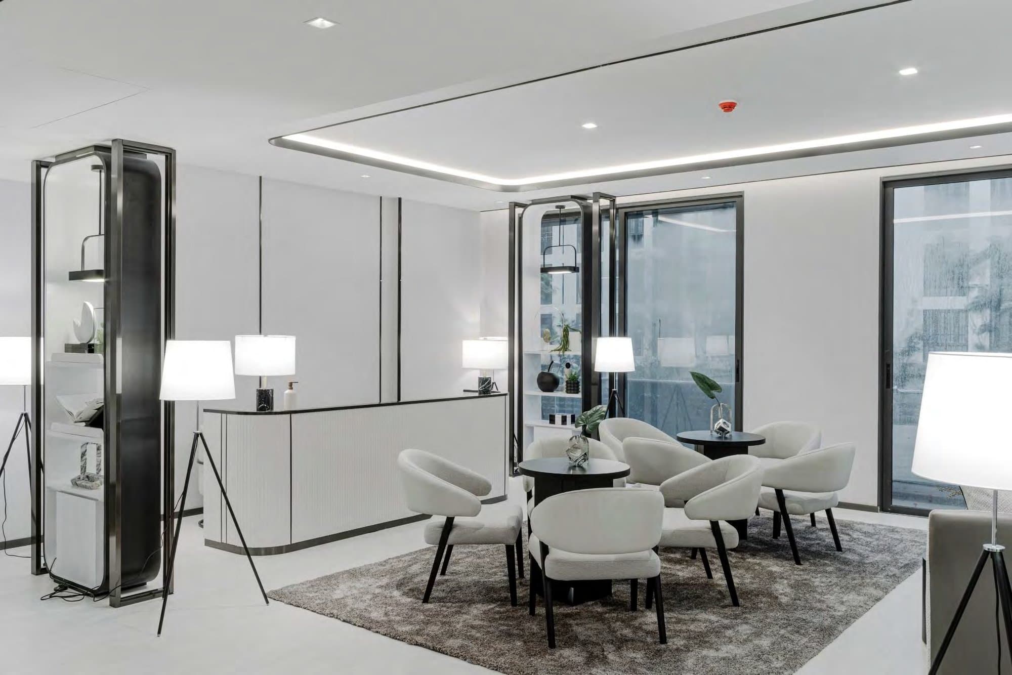 Bright social lounge with white bouclé armchairs, black bistro tables, tripod floor lamps and metal-framed display vitrines on pale flooring.
