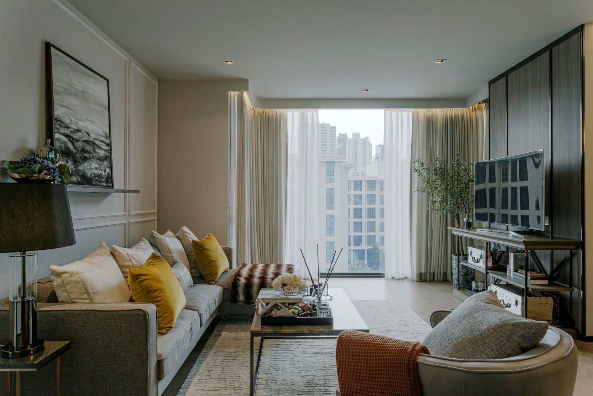 Living room dressed with mustard velvet cushions, a moody seascape canvas, a sheer-curtained window framing a city view and a dark media wall.