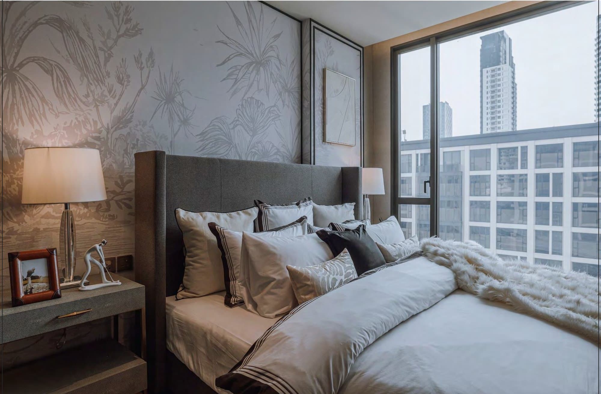 Bedroom with a grey upholstered headboard, layered white and chocolate bedlinen, botanical wallpaper and a floor-to-ceiling window over the city.