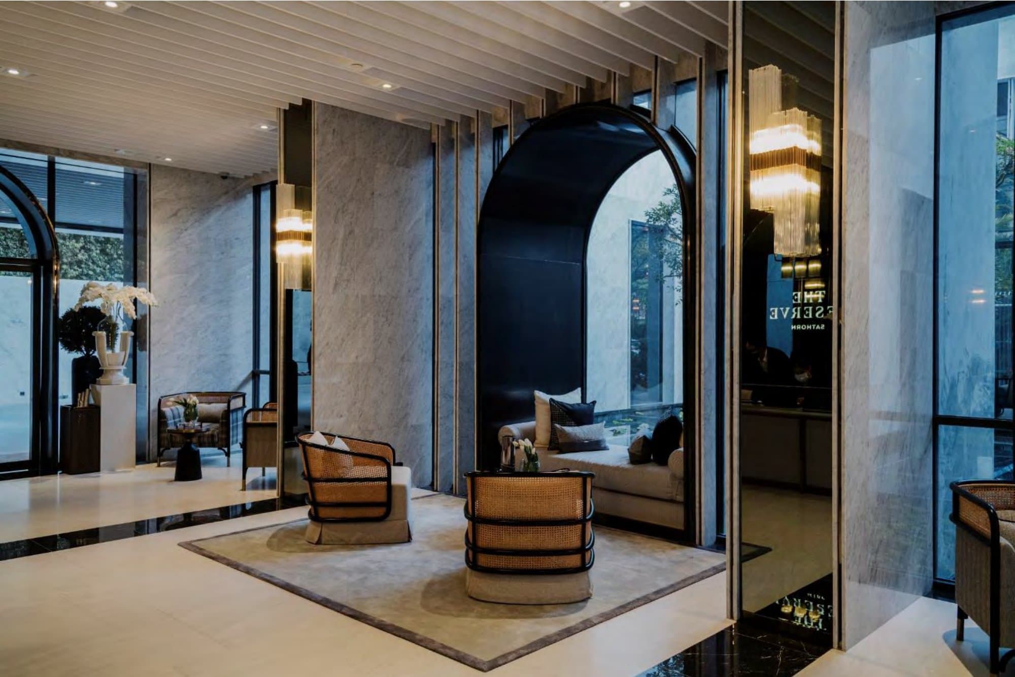 Marble-lined lounge where cane-backed armchairs sit on a soft rug before a glossy black arch opening to a garden view