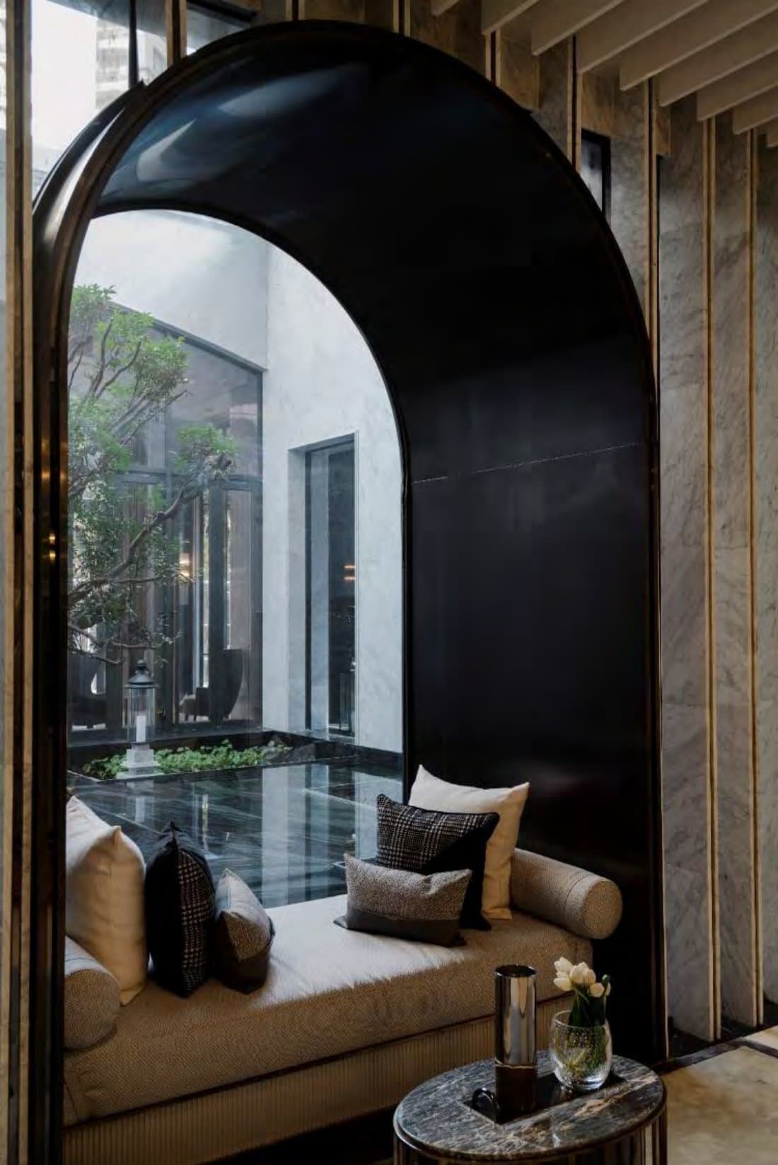 Cushioned daybed set within a tall black arch, overlooking a courtyard tree and mirror-still water garden beyond floor-to-ceiling glass