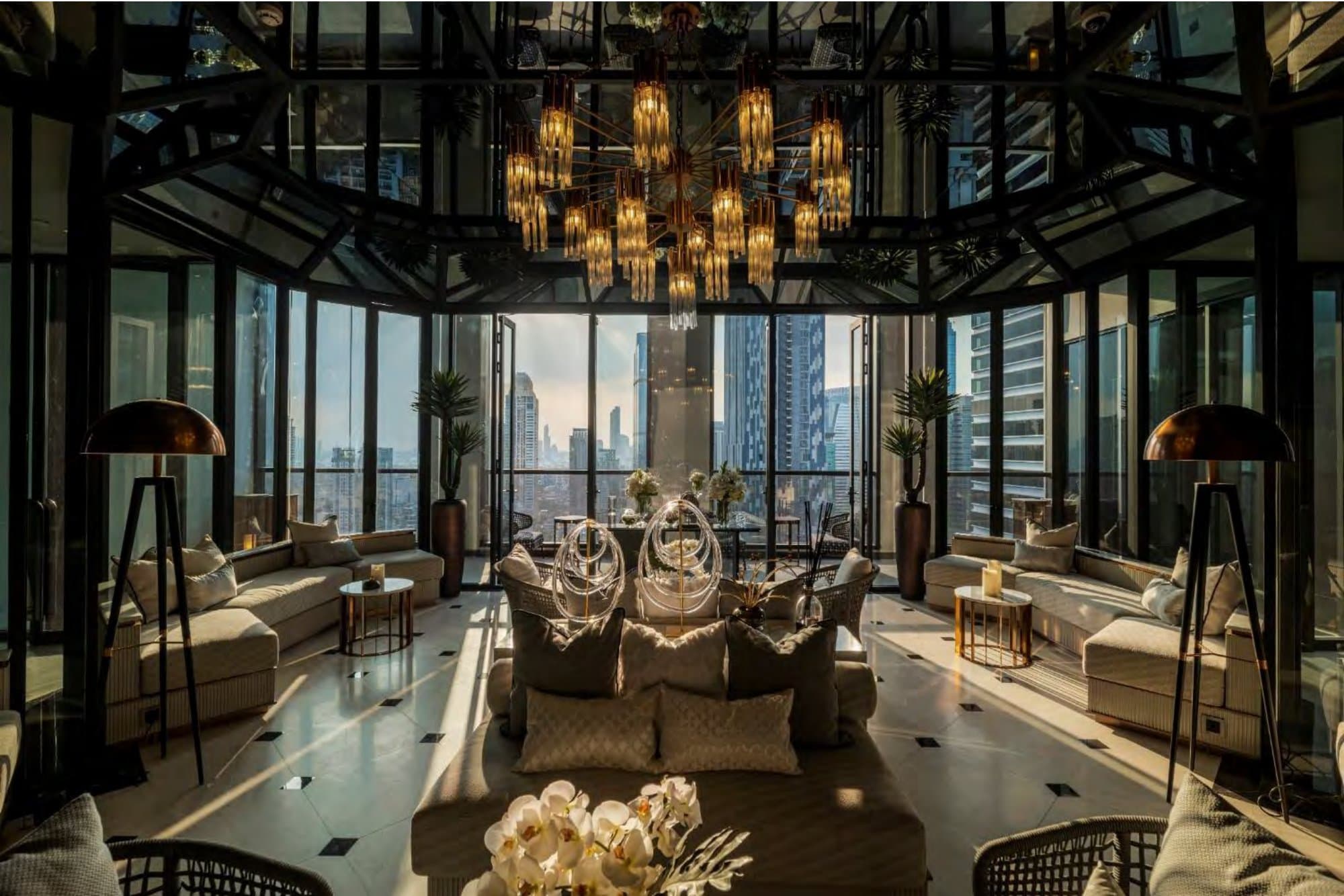 Double-height sky lounge with a cascading crystal chandelier, pale sofas, tripod floor lamps and the Bangkok skyline through faceted glass walls