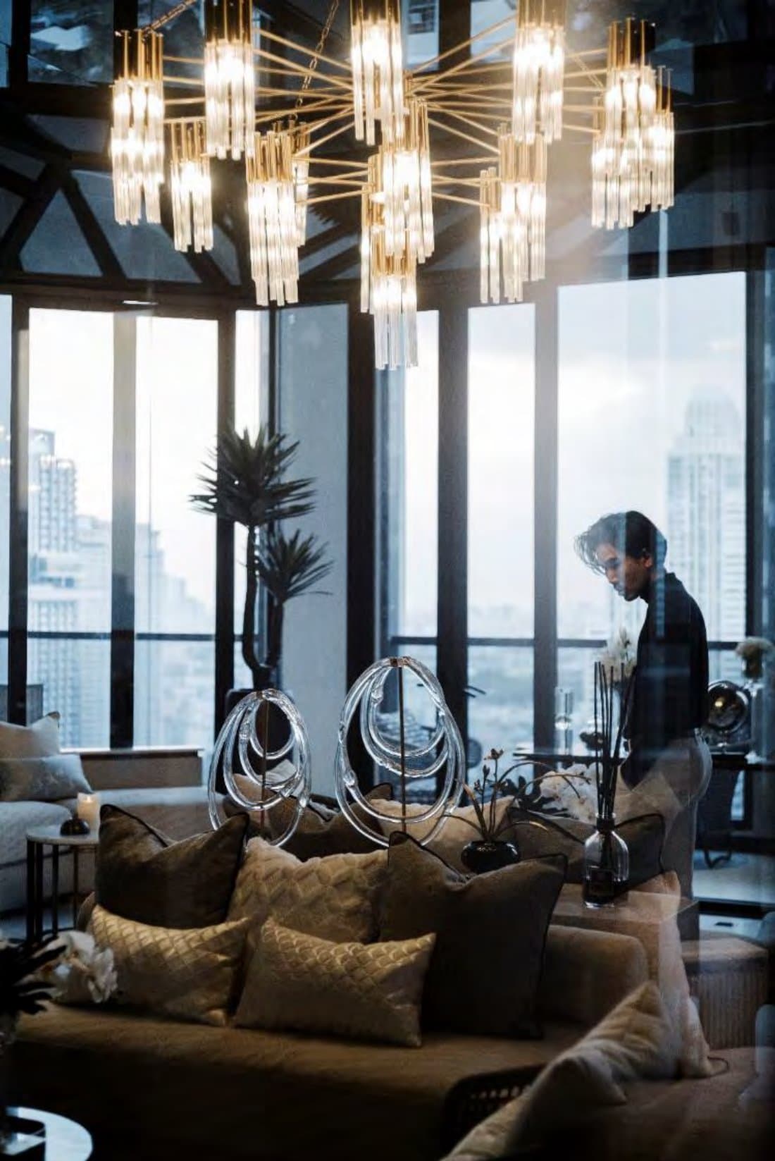Chandelier-lit lounge with layered cushions, twin coiled-glass sculptures and tall windows framing high-rise towers at dusk