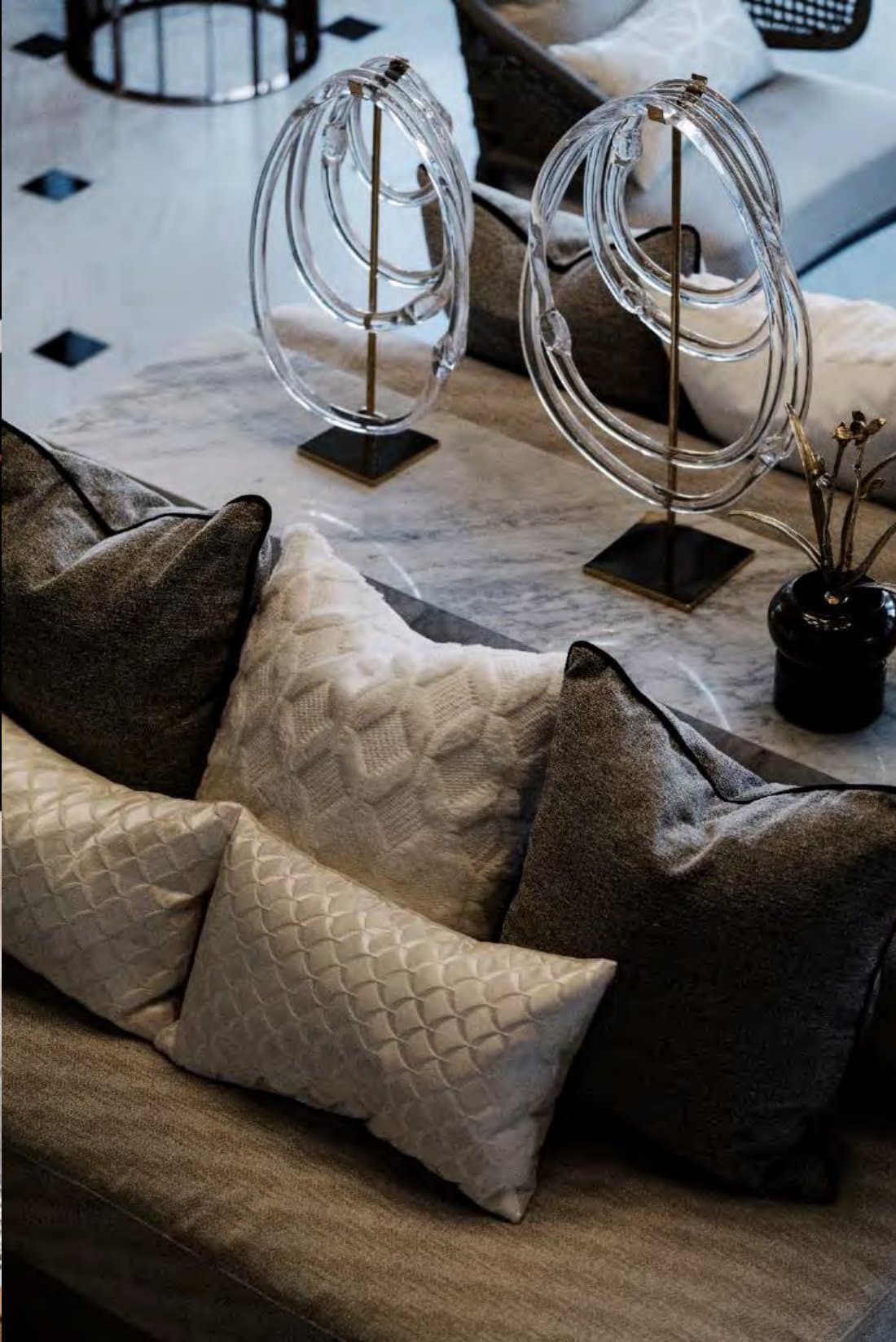 Styling detail of a sofa layered with quilted cream and charcoal cushions beneath two coiled-glass sculptures on brass stands