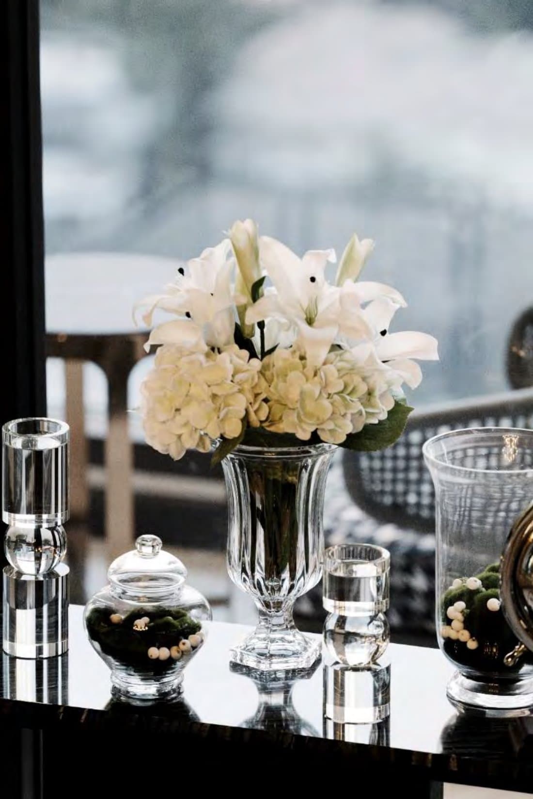 Console vignette of white lilies and hydrangea in a crystal urn, flanked by clear glass objects against a bright window