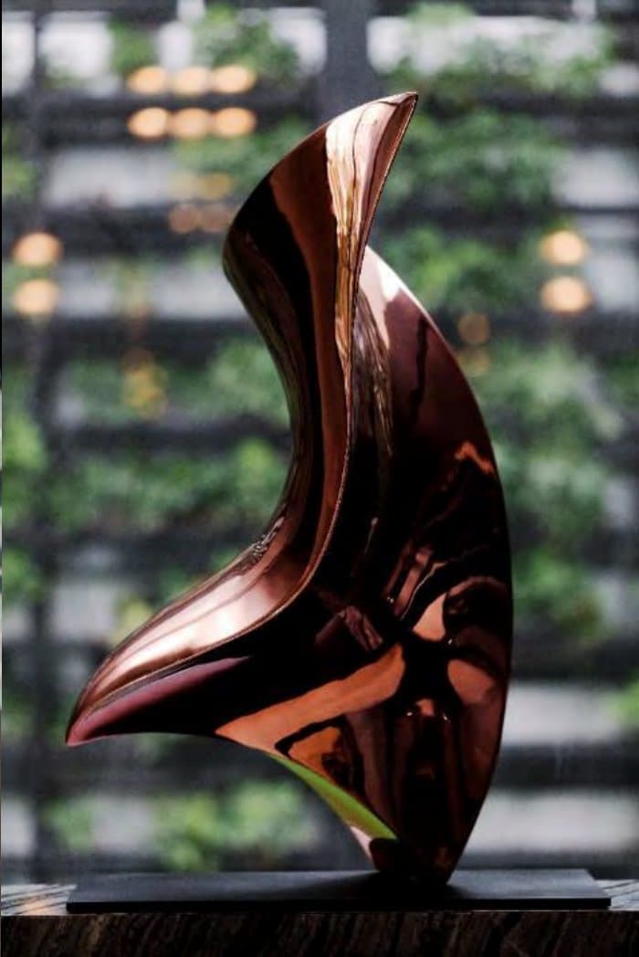 Polished copper abstract sculpture curving upward on a dark plinth, set against a softly blurred green living wall