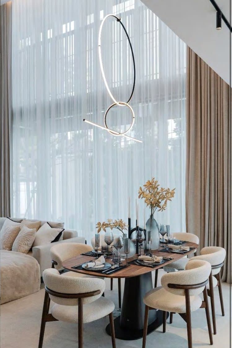 Dining vignette beneath a sculptural looping pendant, walnut table styled with dried stems and boucle armchairs against tall sheer curtains.